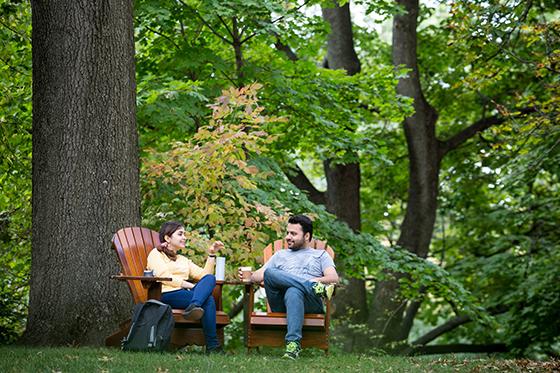 图为两名立博体育官网的学生坐在外面绿草地上高大的橡树下的阿迪朗达克椅子上喝咖啡. 